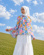A woman wearing a white hijab, a colorful floral long-sleeve blouse, and a long white skirt stands on a grassy hill under a blue sky with clouds.