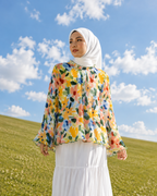 A woman wearing a white hijab, a white tiered maxi skirt, and a colorful floral long-sleeve blouse stands on a grassy hill under a blue sky with clouds.
