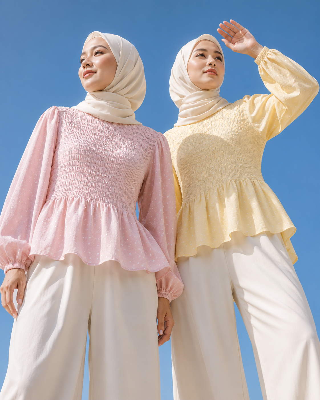 Two women wearing hijabs and adrianna yariqa long-sleeve peplum tops, one in light pink and one in pale yellow, paired with white wide-leg pants, photographed against a clear blue sky.